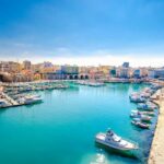 Heraklion port with boats, a promenade, and city buildings under a blue sky.
