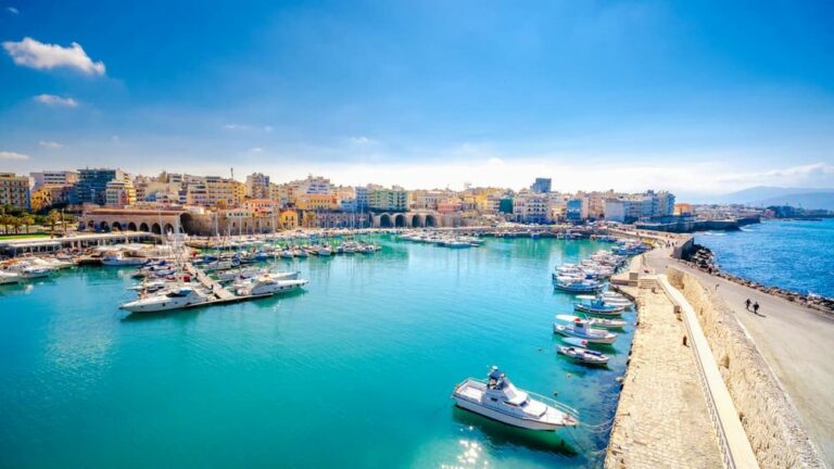 Heraklion port with boats, a promenade, and city buildings under a blue sky. best hotels, villas, and apartments