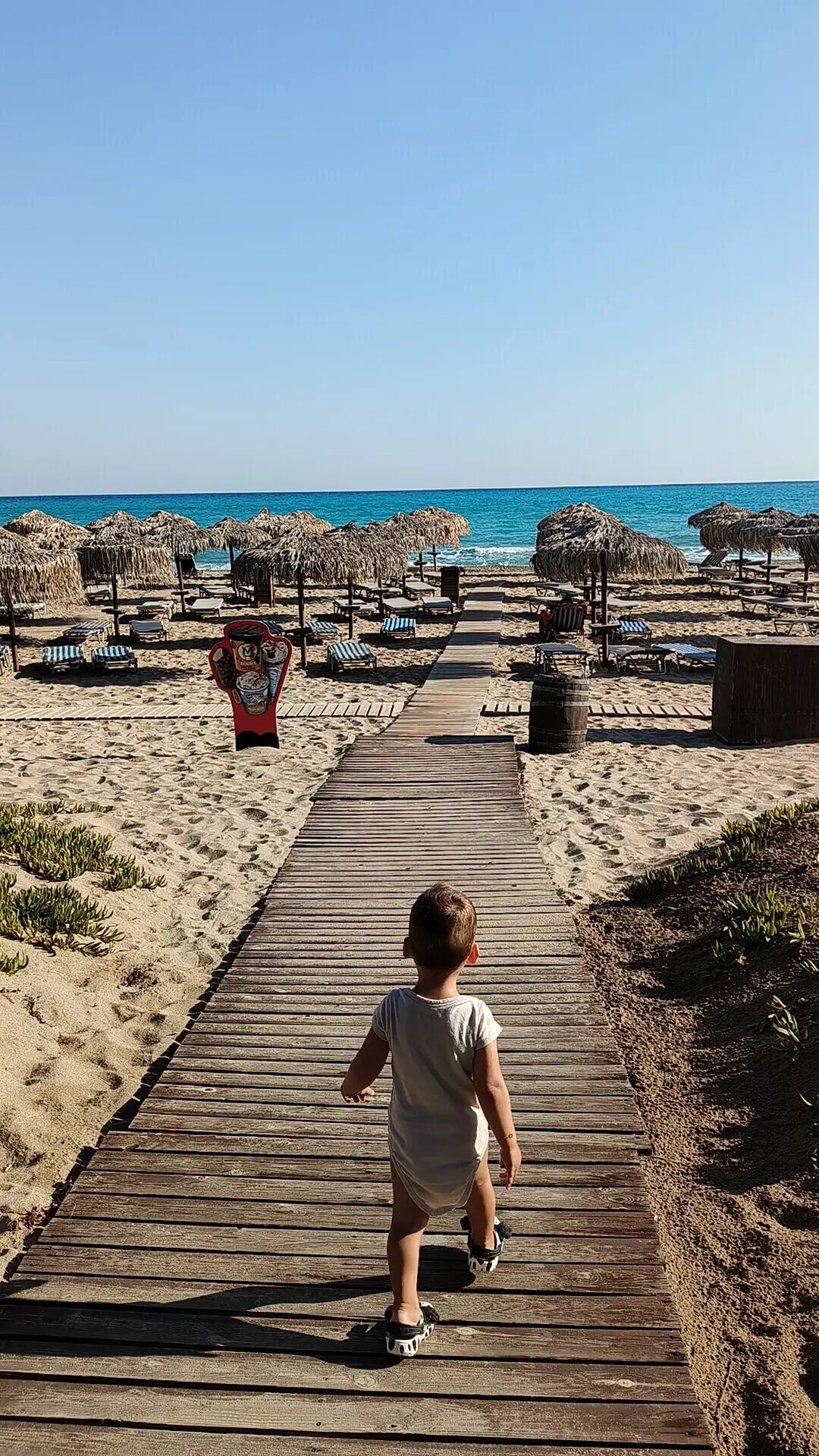 A young child walks along a wooden path leading to a sandy beach in Crete. The beach is adorned with straw umbrellas, sunbeds, and the vibrant blue waters of the Mediterranean under a bright, clear sky.