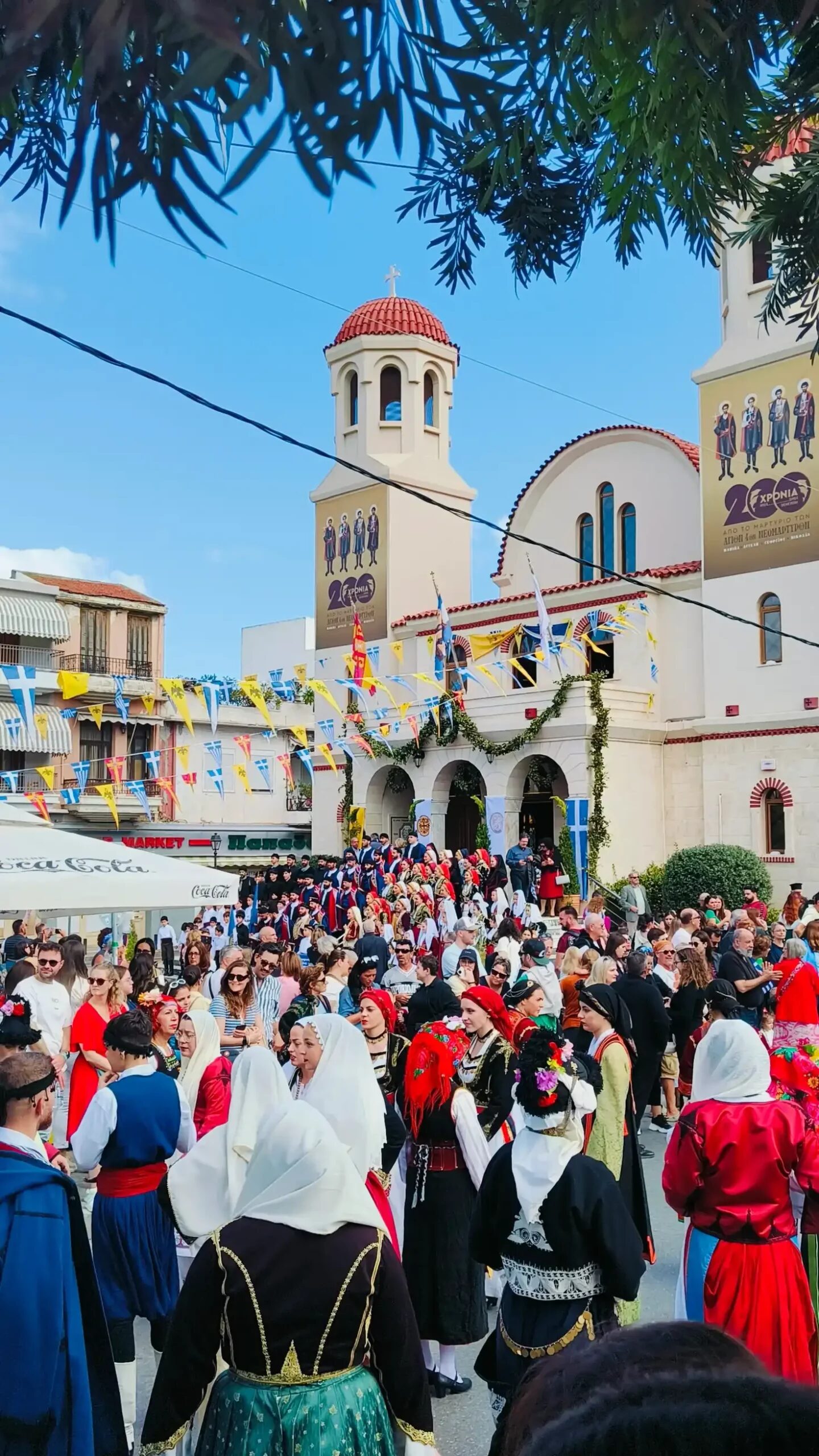 A vibrant scene from the Panigiria festival in Crete, featuring a crowd dressed in traditional Greek attire gathered outside a church decorated with colorful flags and banners under a bright blue sky. The event showcases local life in Crete