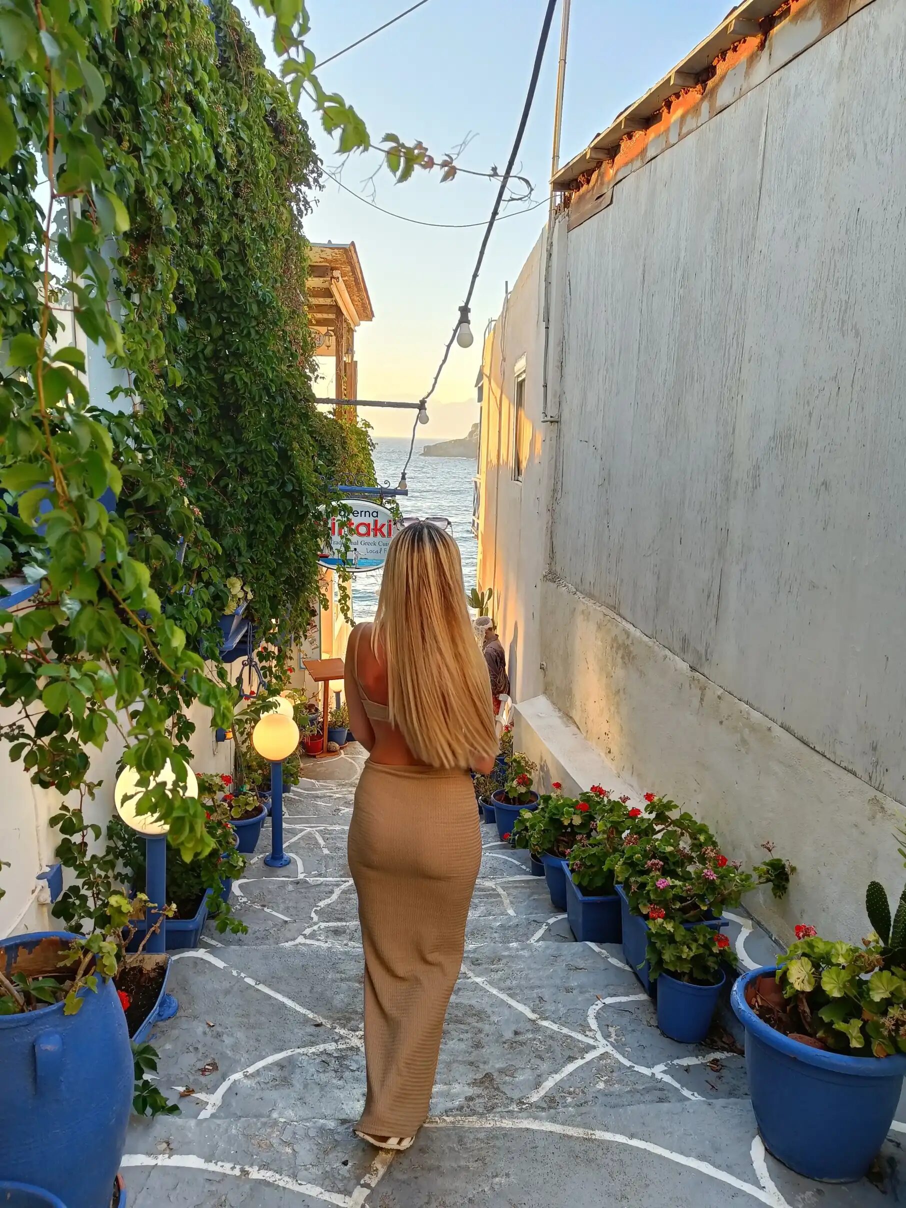 A woman with long blonde hair wearing a beige dress walks down a narrow, charming street in Matala, Crete. The pathway is lined with vibrant flowers in blue pots, and the view opens up to the sea under the warm glow of the sunset.